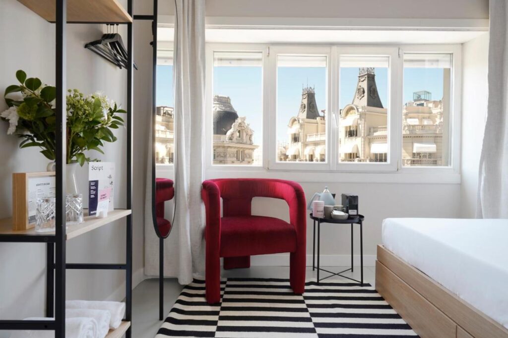 Where to Stay in Madrid: 14 Best Hotels 29 A modern hotel room—perfect for those wondering where to stay in Madrid first time—features a red chair, side table, open shelving, and a black-and-white striped rug. Large windows reveal ornate historic buildings and vibrant city views.
