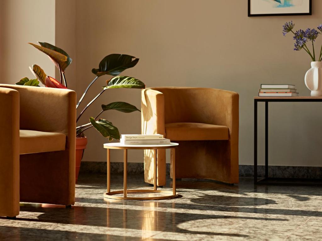 Boutique Hotels in Berlin Kreuzberg 19 A cozy, sunlit sitting area reminiscent of boutique hotels in Berlin Kreuzberg features two tan armchairs, a round coffee table with books, a potted plant, and a side table with stacked books and a white vase of purple flowers. Marble floors reflect the light.