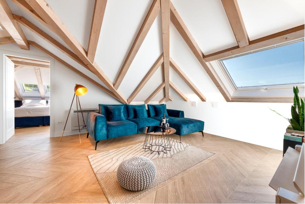 Most Unique Boutique Hotels in Dubrovnik 8 A modern attic living room with exposed wooden beams, a blue velvet sofa, round coffee tables, and a pouf on a beige rug evokes the chic style of boutique hotels in Dubrovnik. Large skylight and doorway to a bright bedroom are visible.