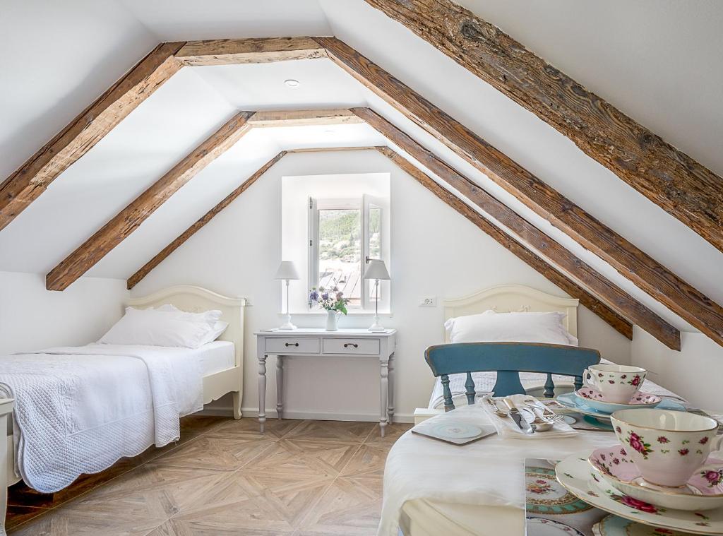 Most Unique Boutique Hotels in Dubrovnik 13 A cozy attic bedroom inspired by boutique hotels in Dubrovnik, with exposed wooden beams, two white single beds, a desk with lamps, and a table set with floral teacups. Sunlight streams in through the window at the far end.
