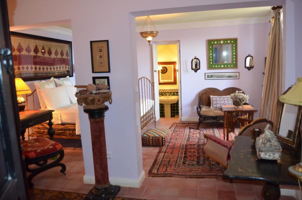 9 UNIQUE Riads in Tangier 29 A cozy, ornately decorated bedroom inspired by riads in Tangier, with antique furniture, patterned rugs, plush cushions, and a small bathtub visible through an open doorway—all set in a softly lit room with pastel walls.