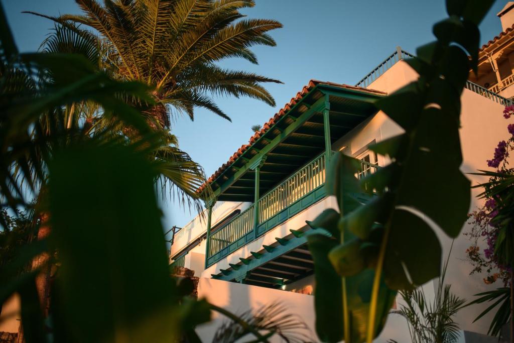 11 UNIQUE Boutique Hotels in Tenerife 29 A white building with green wooden balconies and railings, part of the charming boutique hotels in Tenerife, is partially obscured by lush palm trees and tropical plants, bathed in warm sunlight.