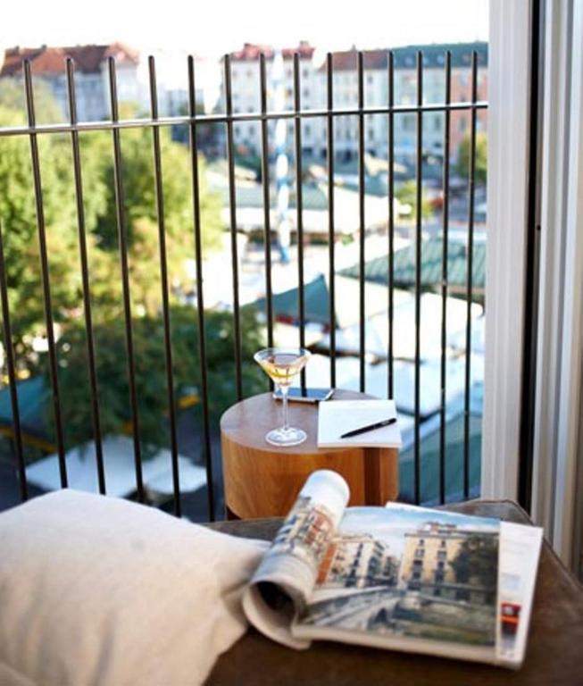 Authentic Boutique Hotels in Munich 14 A magazine and pillow rest on a chair beside a small round table holding a drink and notebook, evoking the cozy charm of boutique hotels in Munich. The scene overlooks a city view through black railings, with buildings and trees in the background.