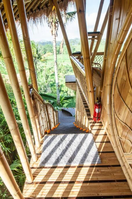 A bamboo staircase with curved railings and a thatched roof leads down to a lush green landscape in one of the best family villas in Bali. Sunlight creates striped shadows on the wooden steps, and a red fire extinguisher is mounted on the wall.