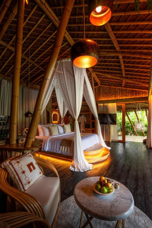 13 Most UNIQUE Places to Stay in Ubud 27 A cozy tropical bedroom with a canopy bed draped in white fabric, bamboo ceiling, warm lighting, and a marble table with fruit—one of the unique places to stay in Ubud—opening to lush greenery outside.