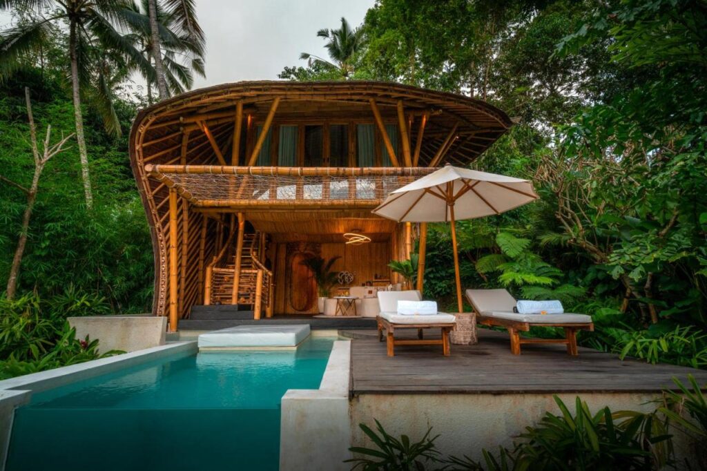 13 Most UNIQUE Places to Stay in Ubud 29 A tropical bamboo villa with a thatched roof, surrounded by lush greenery. Among the unique places to stay in Ubud, it offers a private pool, sun loungers with towels, and a white umbrella on a wooden deck for a relaxing outdoor retreat.