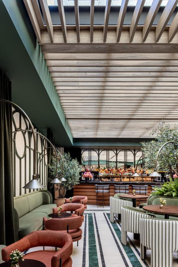 Where to Stay in Madrid: 14 Best Hotels 54 A stylish restaurant interior with green and terracotta chairs, striped sofas, lush plants, and a wooden bar—perfect for those searching where to stay in Madrid first time. Natural light from a slatted ceiling creates a modern, inviting atmosphere.