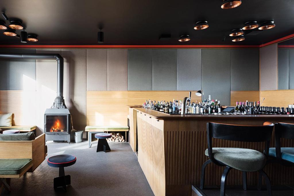 Authentic Boutique Hotels in Munich 6 A modern bar in the style of boutique hotels in Munich, featuring a wooden counter, shelves of bottles, green cushioned seating, a black freestanding fireplace with a blazing fire, and minimalist lighting on a dark ceiling.