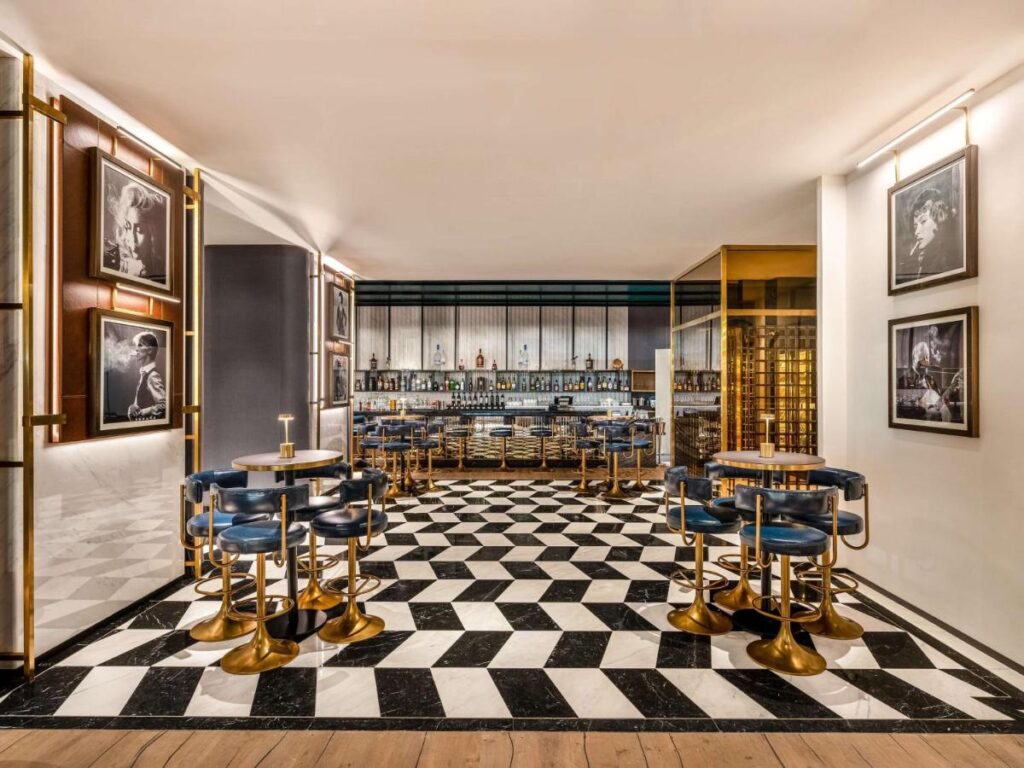 Where to Stay in Doha, Qatar: Transit, City & Beach Hotels 26 A stylish bar interior with geometric black-and-white floor tiles, blue cushioned chairs with gold frames, and round tables—a chic spot often recommended for those seeking where to stay in Doha, complete with framed photos and a well-stocked bar.