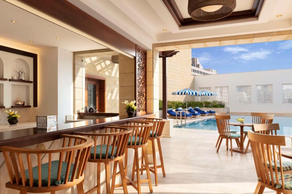 Where to Stay in Doha, Qatar: Transit, City & Beach Hotels 41 A bright, modern cafe with wooden chairs and tables overlooks an outdoor pool with blue lounge chairs and striped umbrellas—an inviting spot for those seeking where to stay in Doha under a sunny sky.