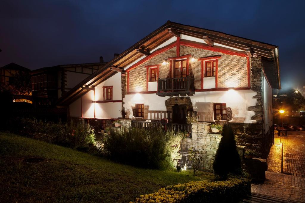 8 Unique Boutique Hotels in Bilbao Spain 29 A rustic, two-story house with stone and white stucco walls, warmly lit at night. Reminiscent of boutique hotels in Bilbao, it features wooden beams, red-trimmed windows, a small balcony, and a garden with grass and bushes.