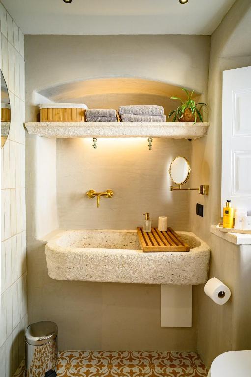 Most Unique Boutique Hotels in Dubrovnik 15 A stylish bathroom inspired by boutique hotels in Dubrovnik, featuring a large stone sink, golden faucet, and a wooden tray. Above the sink are folded towels and a small potted plant on a shelf. Patterned tiles adorn the light-colored walls and floor.