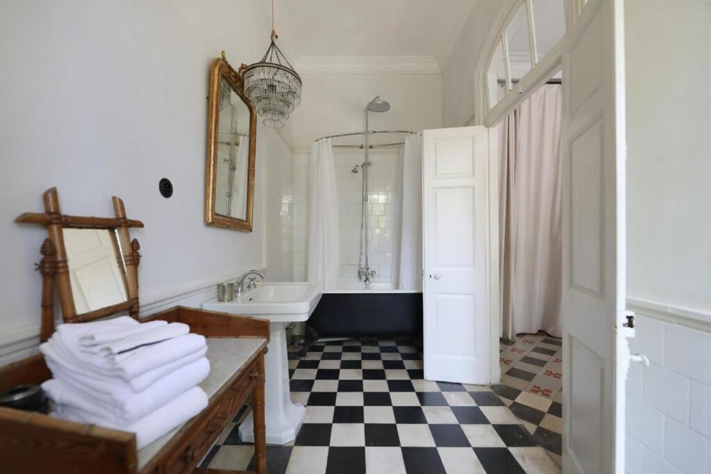 9 UNIQUE Riads in Tangier 4 A bright bathroom inspired by riads in Tangier features a black-and-white checkered floor, pedestal sink, vintage mirror, chandelier, freestanding bathtub with shower, and a wooden shelf holding neatly folded white towels.