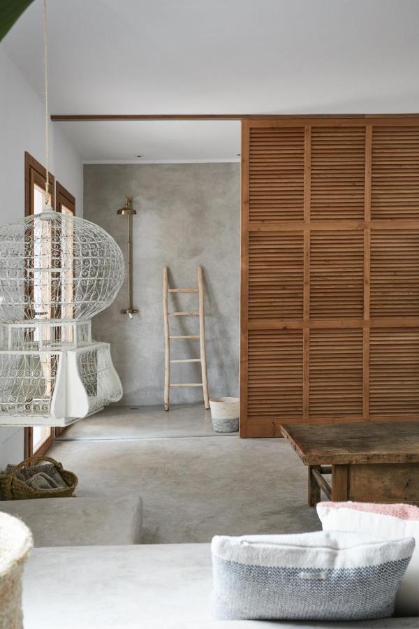 A modern, minimalist living space inspired by boutique hotels in Formentera, featuring a wooden slat sliding door, gray concrete floors, a rattan hanging chair, neutral-toned pillows, a wooden ladder, and an open shower with a brass fixture.