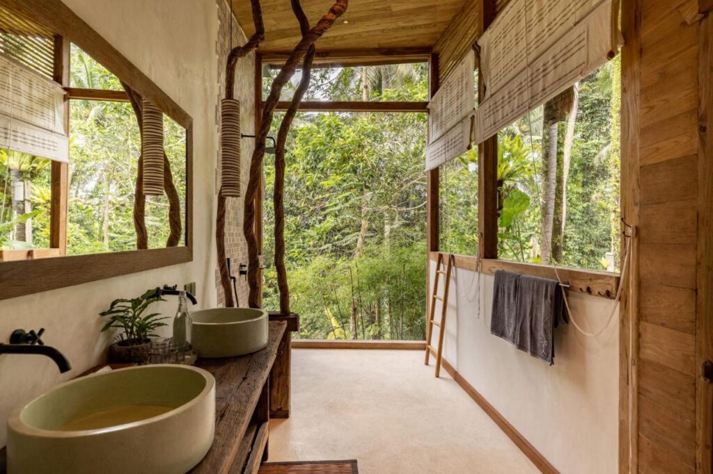 A rustic bathroom in one of the best family villas in Bali, with large windows framing lush greenery. It features two round sinks on a wooden counter, a towel on a wooden ladder, and natural wood and stone accents throughout.