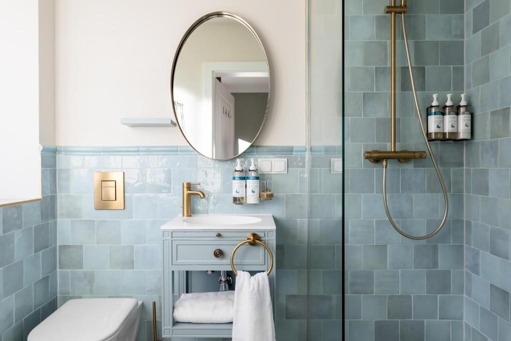 Unique Boutique Hotels in Salzburg 17 A modern bathroom inspired by boutique hotels in Salzburg, featuring blue-green tiled walls, a round mirror above a small sink with gold fixtures, a towel below, a glass shower with gold hardware, and toiletries on a glass shelf.