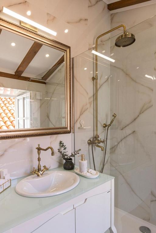 Most Unique Boutique Hotels in Dubrovnik 28 A stylish bathroom reminiscent of boutique hotels in Dubrovnik features a white vanity, gold fixtures, a large mirror, and marble-patterned walls. A small plant and toiletries rest on the counter beneath exposed wooden ceiling beams.