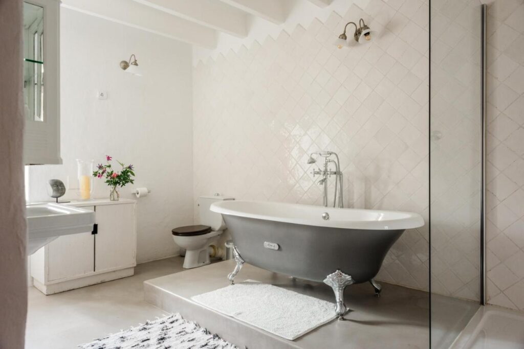 10 AUTHENTIC Finca Hotels in Ibiza 24 A bright bathroom, inspired by finca hotels in Ibiza, features a black clawfoot bathtub on a raised platform, white tiled walls, a white rug, wooden cabinet with flowers, wall lamp, and a modern shower with glass partition.