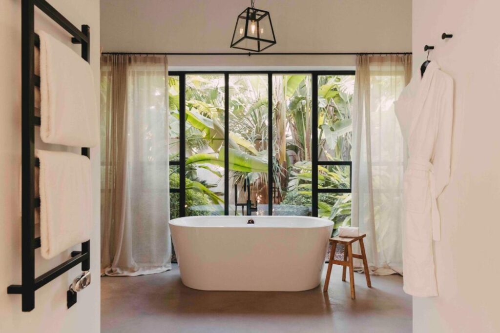 Unique Agroturismos in Mallorca 32 A modern bathroom in the style of agroturismos in Mallorca, featuring a freestanding white bathtub, large glass windows with lush greenery outside, sheer curtains, a wooden stool, towel rack with towels, and a hanging bathrobe.