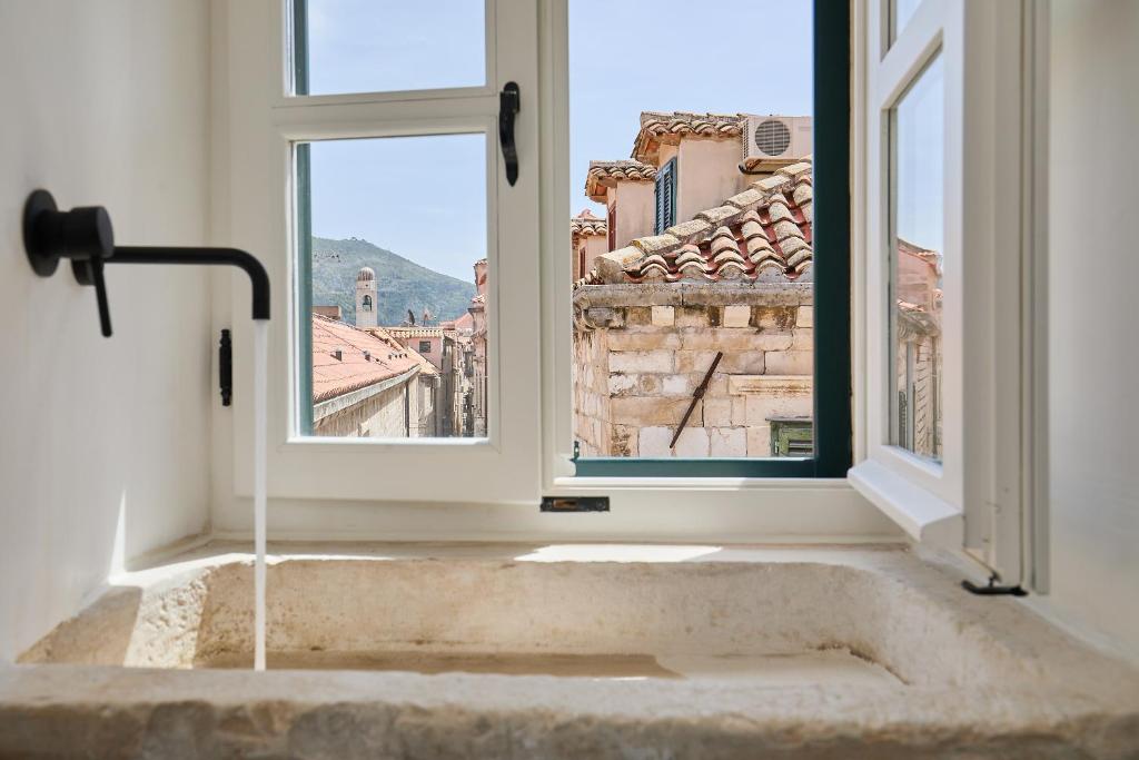 Most Unique Boutique Hotels in Dubrovnik 23 A stone sink with running water sits beneath an open window in one of the boutique hotels in Dubrovnik, offering a view of terracotta rooftops and old stone buildings, with hills visible in the background under a clear sky.