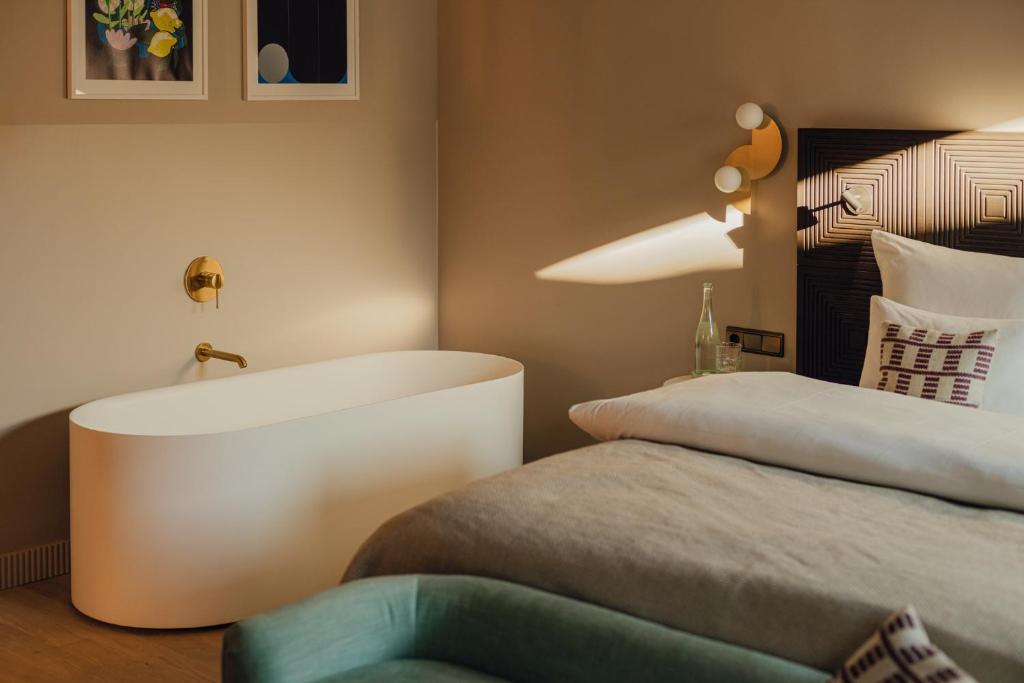 Unique Boutique Hotels in Salzburg 6 Modern bedroom reminiscent of boutique hotels in Salzburg, featuring a freestanding white bathtub with a gold faucet, neatly made bed with patterned pillows, green sofa, wall-mounted bedside light, and framed art on beige walls. Sunlight streams through the room.