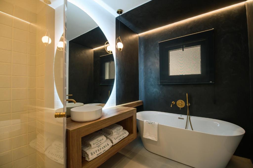 11 UNIQUE Boutique Hotels in Tenerife 31 Modern bathroom inspired by boutique hotels in Tenerife, featuring a freestanding white bathtub, black walls, a round illuminated mirror, wooden vanity with towels, vessel sink, gold fixtures, and a frosted window letting in soft light.