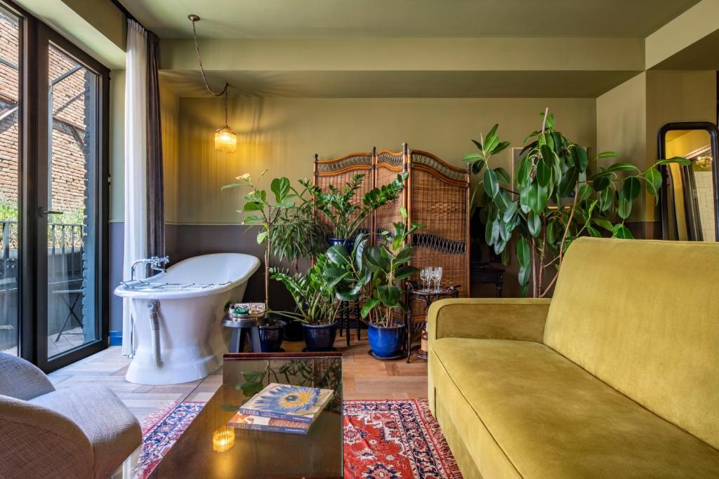 11 Unique Hotels in Georgia You Can't Miss! 29 A cozy living room, inspired by unique hotels in Georgia, features a yellow sofa, a glass coffee table with books, a vintage rug, many potted plants, a freestanding bathtub near large windows, and a folding screen in the background.