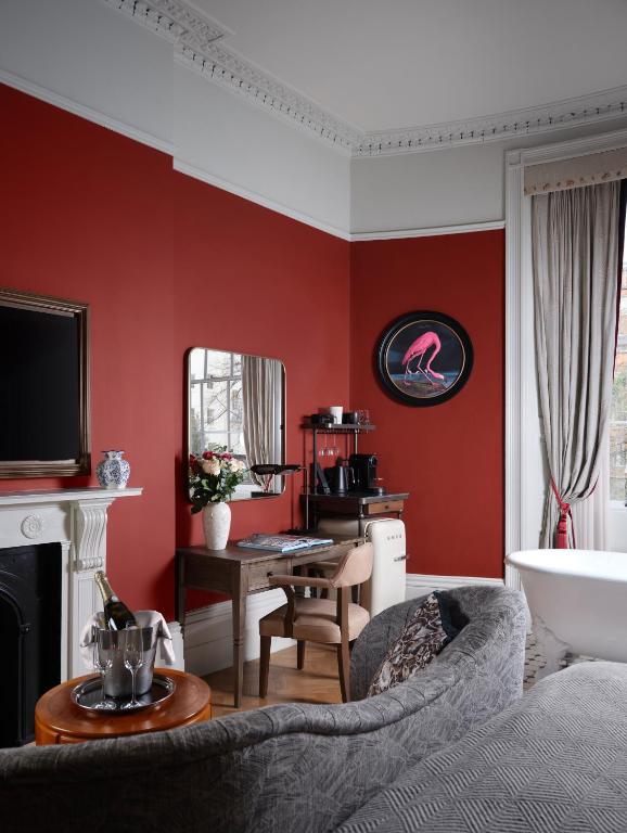 15 Best Boutique Hotels in London, UK 33 A stylish room inspired by boutique hotels in London, featuring red walls, a fireplace, a TV, a desk with a mirror, a coffee station, flamingo wall art, a gray couch, wine on a round side table, and a freestanding bathtub by tall curtained windows.