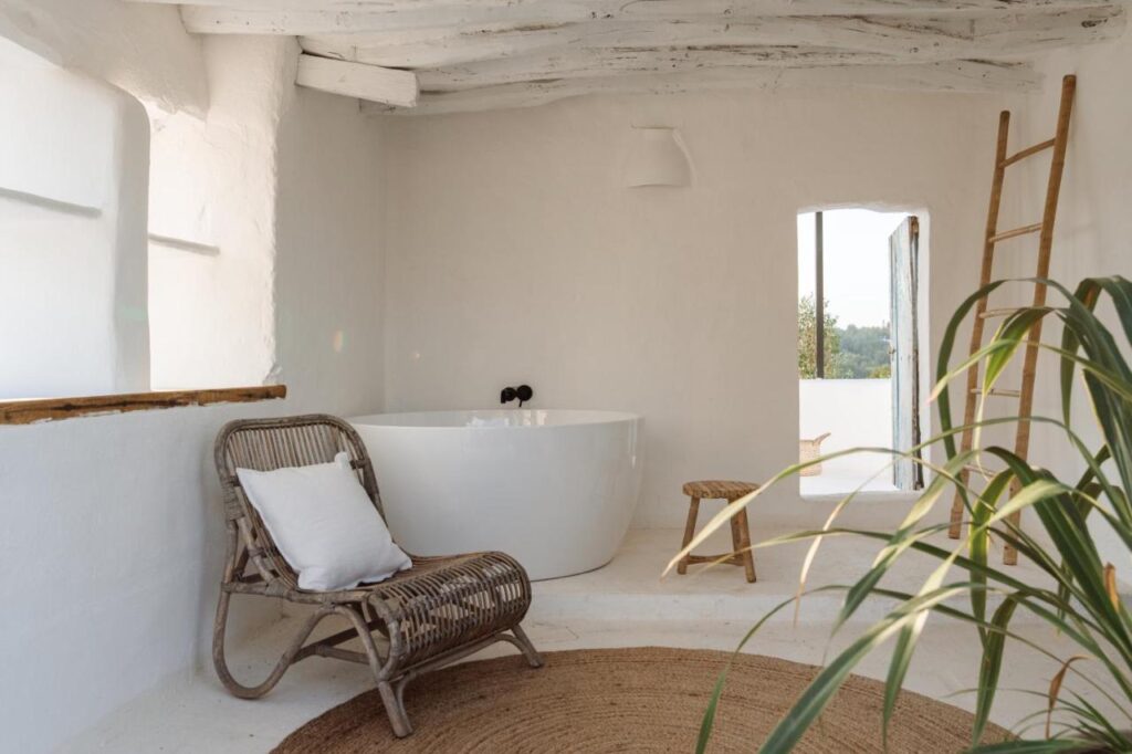 10 AUTHENTIC Finca Hotels in Ibiza 4 A bright, minimalist bathroom inspired by finca hotels in Ibiza features white walls, a freestanding bathtub, a wooden chair with a white cushion, a round jute rug, a wooden stool, a ladder, and a plant by the arched window.