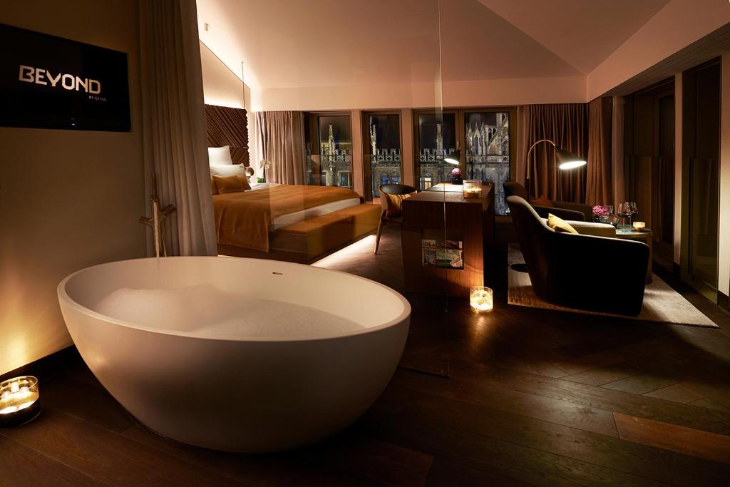 Authentic Boutique Hotels in Munich 33 A luxurious hotel suite at one of the finest boutique hotels in Munich features a modern freestanding bathtub, a cozy seating area, a bed with warm lighting, large windows with city views, candles, and contemporary décor.