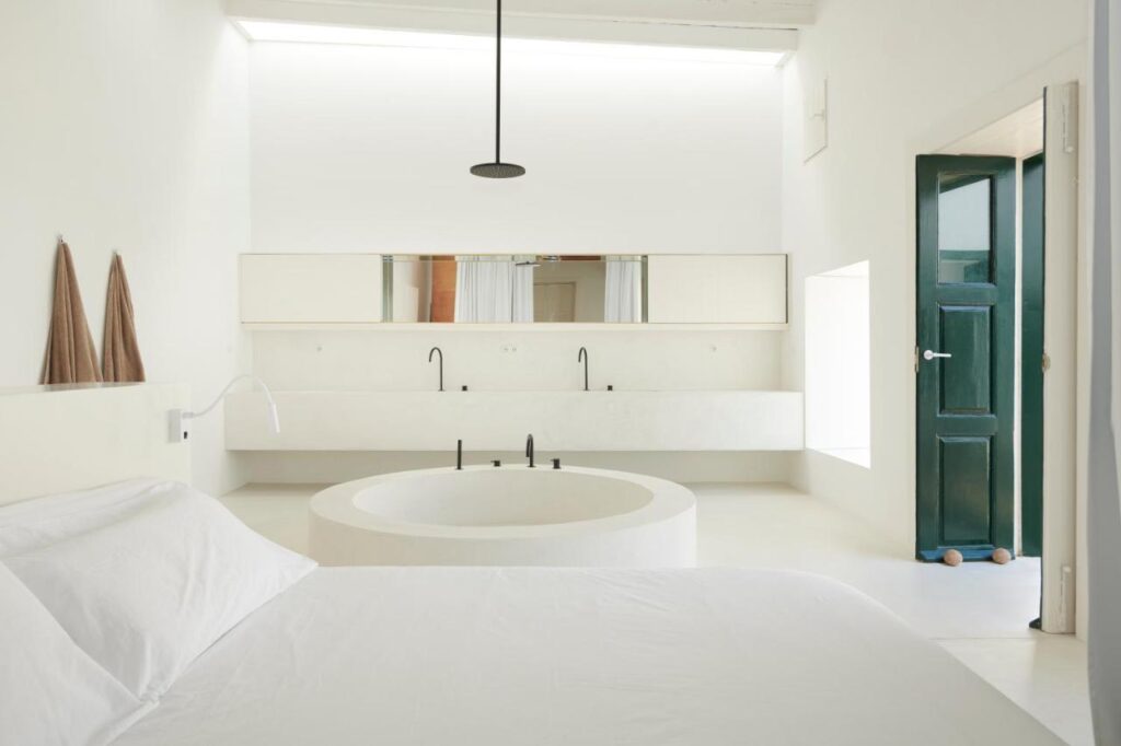Adults Only Hotels in Fuerteventura 4 Minimalist white bedroom with a round sunken bathtub at the foot of the bed, double sinks with black fixtures, two brown towels on the wall, and an open green door letting in natural light—perfect for adults only hotels in Fuerteventura.
