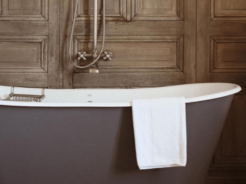 11 Unique Places to Stay in Provence 9 A freestanding bathtub with a white towel draped over the side sits in front of rustic, wooden paneled doors—an inviting scene often found in unique places to stay in Provence. An exposed metal shower fixture is mounted on the wall above the tub.