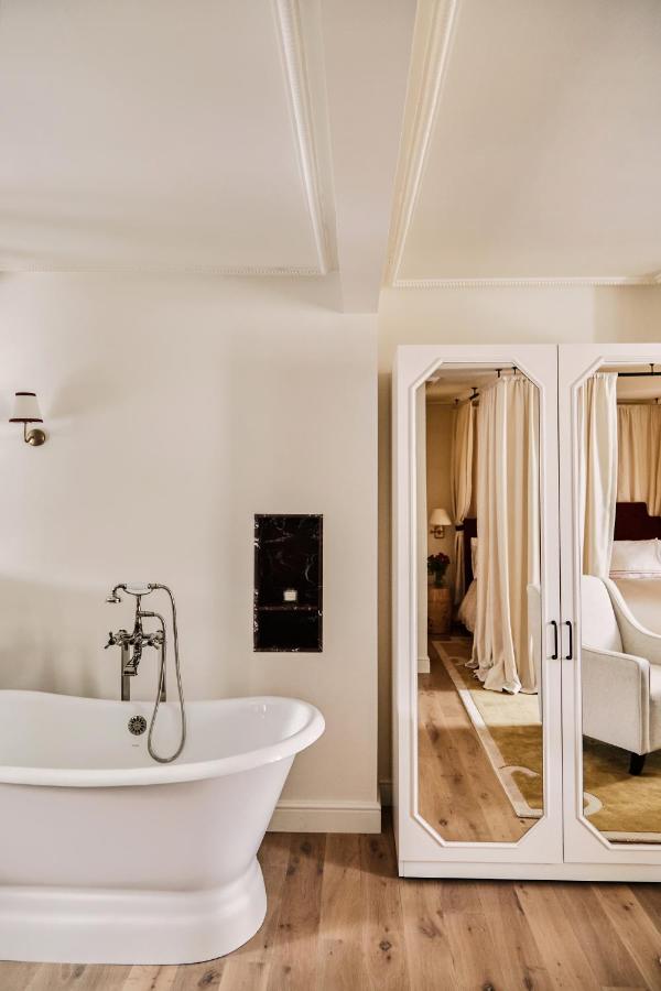 15 Best Boutique Hotels in London, UK 8 A modern bathroom in the style of the best boutique hotels London offers, featuring a white freestanding bathtub, wooden flooring, and a mirrored wardrobe reflecting a cozy bedroom with beige curtains and furnishings.