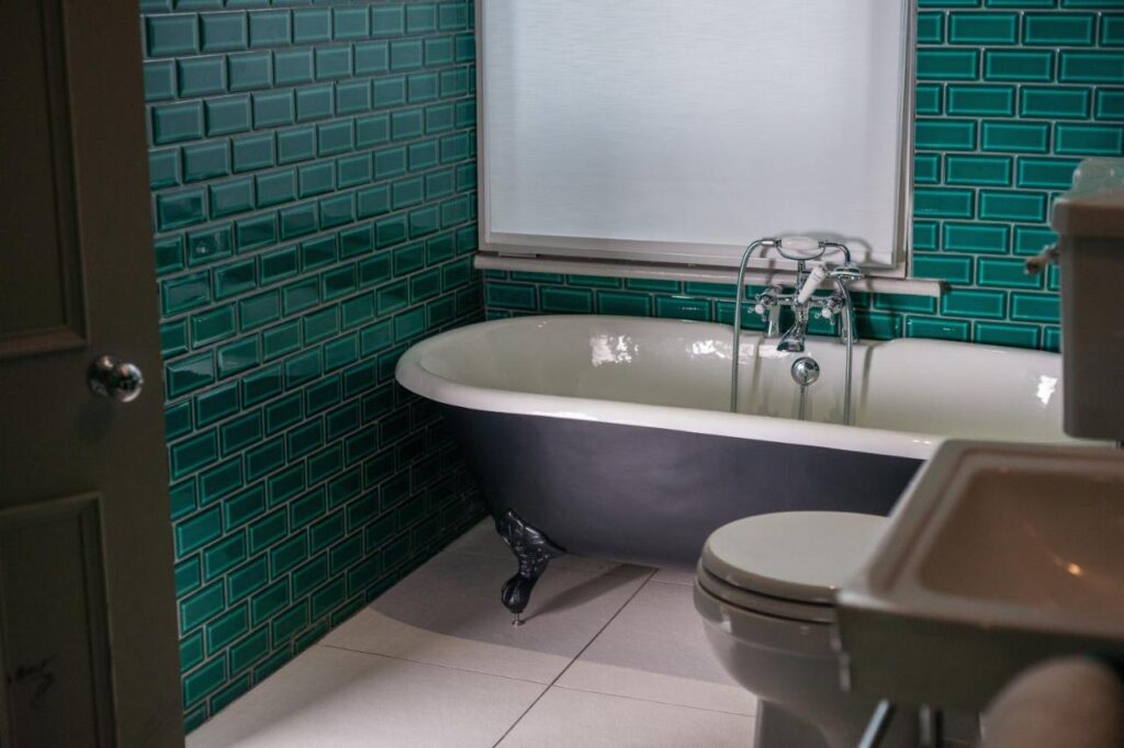 15 Best Boutique Hotels in London, UK 15 A bathroom inspired by the best boutique hotels London offers, featuring teal subway tile walls, a white clawfoot bathtub with black exterior, a toilet, and a white sink. A window above the tub fills the space with natural light.