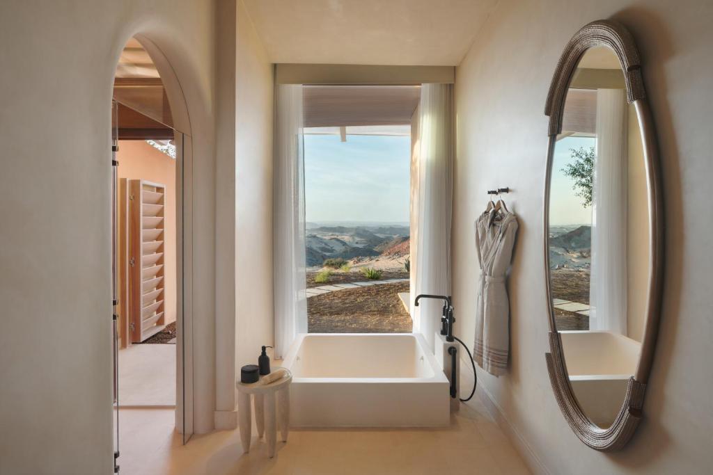 9 Best Hotels in Saudi Arabia: Unique Stays Across the Country 27 Minimalist bathroom inspired by the best hotels in Saudi Arabia, featuring a bathtub under a large window with scenic mountain views, a round mirror, and a robe beside the tub. Soft neutral tones and natural light enhance the tranquil space.