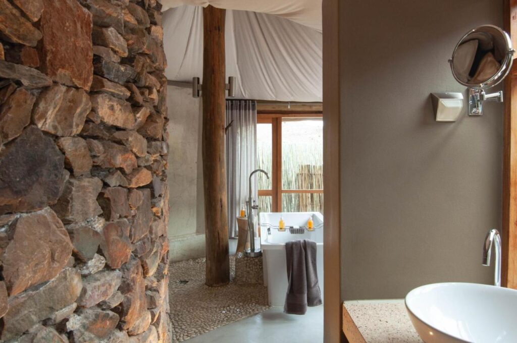 A rustic bathroom reminiscent of Game Lodges in Western Cape features a stone wall, freestanding bathtub with towels, wooden pillars, pebbled shower floor, and a modern sink with round mirror on a beige wall. Natural light streams in through the window.
