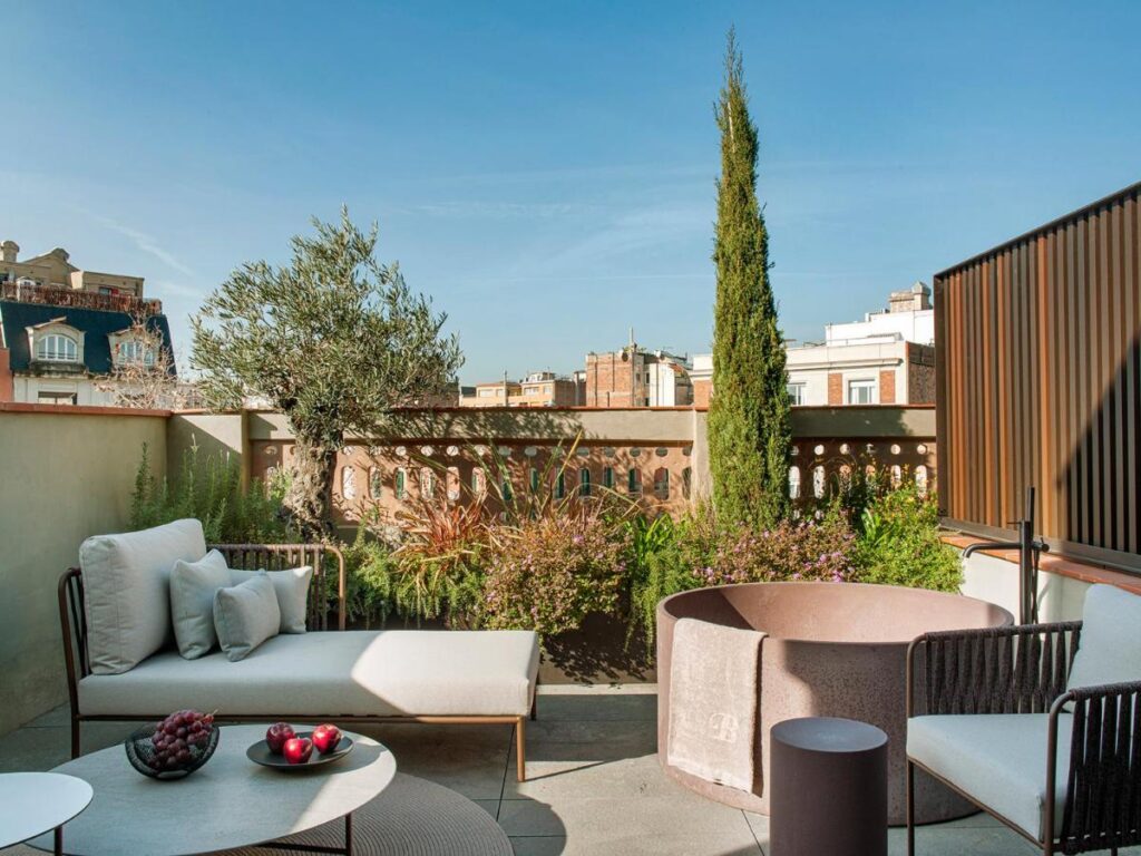 Top Barcelona Hotels with Rooftop Pool 8 A stylish outdoor patio at one of the top Barcelona Hotels with Rooftop Pool features cushioned seating, a round coffee table with fruit, a circular planter, lush greenery, and city buildings framed by a clear blue sky.