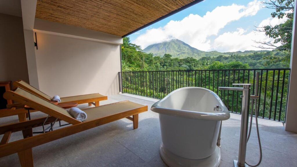 Unique Hotels in Costa Rica: Top Stays 2 A luxurious open-air terrace at one of the unique hotels in Costa Rica features a freestanding bathtub and two wooden loungers, overlooking lush greenery and a distant mountain under a partly cloudy sky.