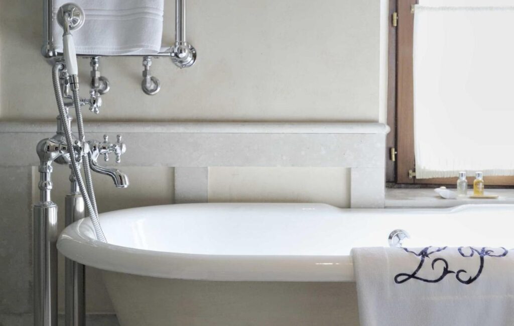 25 Weekend Trips from Madrid Max. 2h Away 56 A white freestanding bathtub with chrome fixtures sits next to a window—perfect for relaxing after weekend trips from Madrid. A towel embroidered with blue letters hangs on the tub, and toiletries are arranged on the bright, clean windowsill.