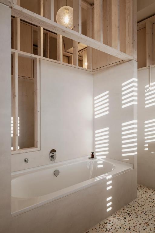 Boutique Hotels in Berlin Kreuzberg 10 A minimalist bathroom reminiscent of boutique hotels in Berlin Kreuzberg, with a white bathtub, light wood framing, and terrazzo floor. Sunlight streams through slatted openings, casting striped shadows across the serene space.