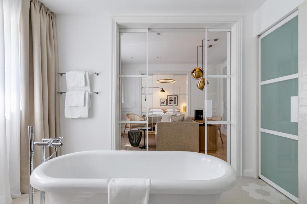 8 TOP Boutique Hotels in Split 10 A modern, white bathroom with a freestanding bathtub, towel rack, and floor-to-ceiling curtains—ideal for those searching Where to Stay in Split. Glass sliding doors reveal a stylish living area with beige chairs and contemporary decor in the background.