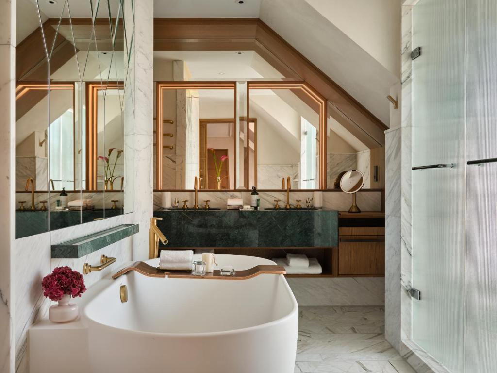 Authentic Boutique Hotels in Munich 28 Inspired by boutique hotels in Munich, this modern bathroom features a freestanding white bathtub, gold fixtures, marble surfaces, large mirrors, a dark green countertop, flowers, and a glass shower enclosure under a sloped ceiling.