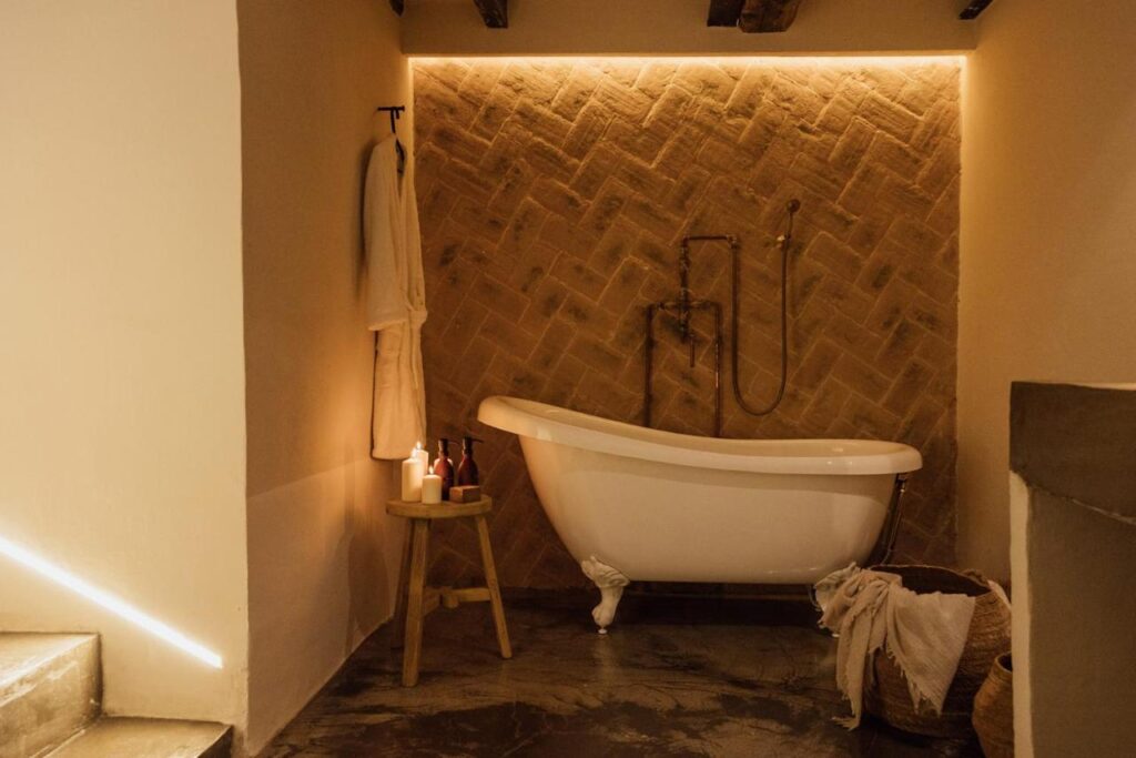 A cozy bathroom, inspired by boutique hotels in Asturias, with a white clawfoot bathtub, warm lighting, a towel and robe on the wall, candles and bottles on a wooden stool, and a woven basket on a stone floor.