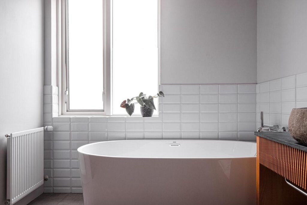 Where to Stay in Reykjavík: Top Guide 25 Minimalist bathroom reminiscent of where to stay in Reykjavik, featuring a white freestanding bathtub, two potted plants on the window sill, white tiled walls, a left-side radiator, and a wooden vanity with a stone sink.