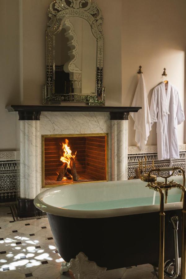 7 UNIQUE Riads in Fez You Won't Forget 15 A luxurious bathroom, inspired by riads in Fez, features a freestanding black-and-white bathtub, a marble fireplace with a lit fire, an ornate mirror above the mantel, and a white bathrobe and towel hanging on the wall.