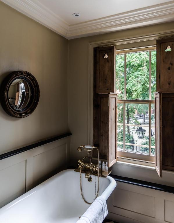 15 Best Boutique Hotels in London, UK 39 A vintage-style bathroom reminiscent of boutique hotels in London, with a white tub, brass faucet, round mirror, and an open wooden-shuttered window overlooking green trees and a street lantern outside.