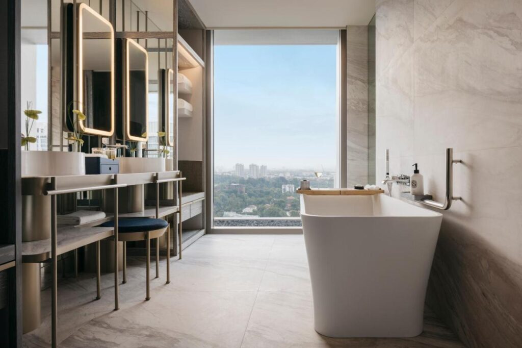 13 Most Unique Hotels in Singapore for an Incredible Holiday 41 Luxurious modern bathroom in one of the unique hotels in Singapore, featuring a freestanding white tub, double vanity with mirrors, marble walls and floors, and a large window offering a cityscape view under a blue sky.