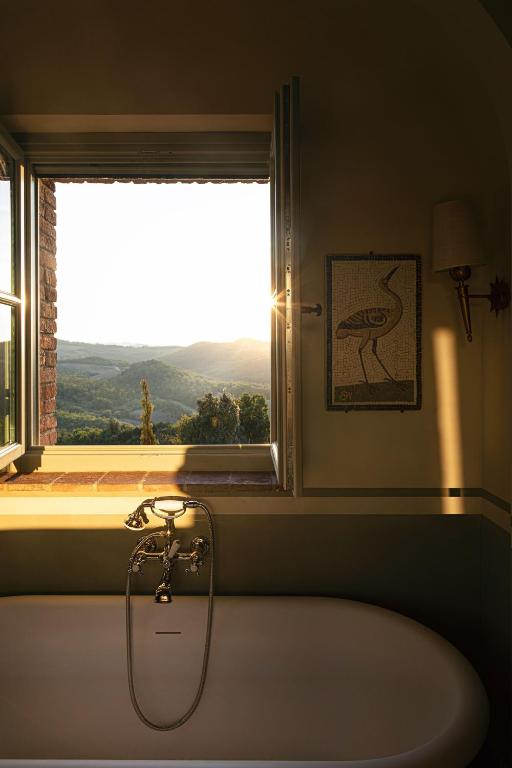 7 Best Agriturismos in Tuscany 15 A bathtub sits beneath an open window with sunlight streaming in, revealing a scenic view of rolling hills and trees at sunset—capturing the tranquil charm of agriturismos in Tuscany. A wall lamp and bird artwork hang beside the window.