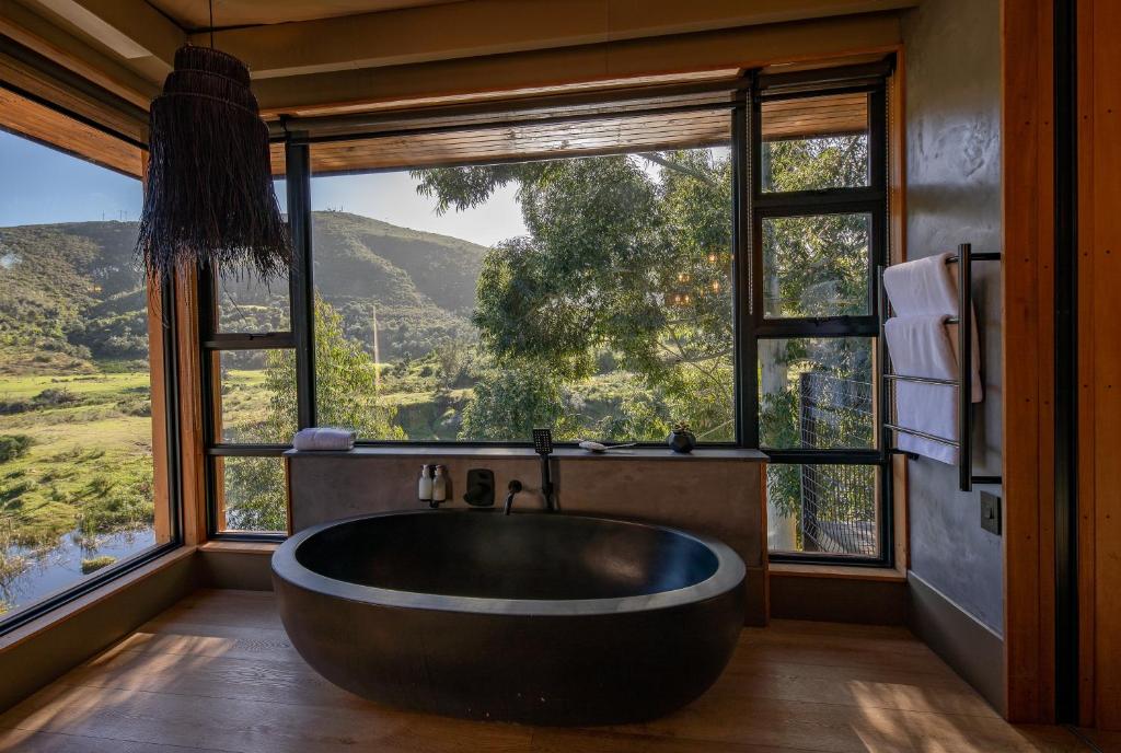 A modern bathroom with a freestanding black oval bathtub in front of large windows, offering scenic views of green hills typical of Game Lodges in Western Cape. Towels hang on a rack to the right, and sunlight fills the cozy space.