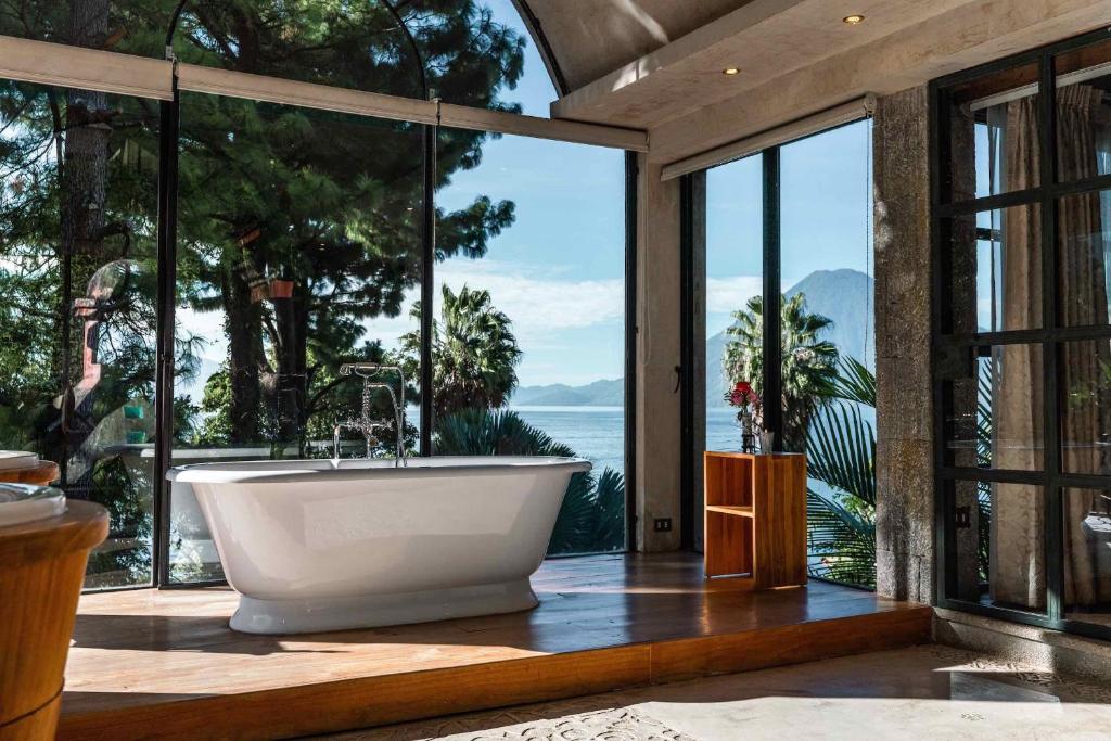 8 UNIQUE Places Where to Stay in Lake Atitlán 3 A modern bathroom with a standalone white bathtub on a wooden platform, surrounded by large glass windows offering a scenic view—perfect inspiration for those searching where to stay in Lake Atitlan.
